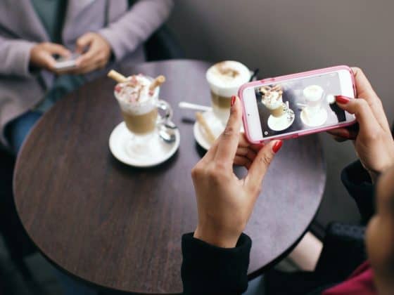An overhead photograph taken from behind a person with red fingernails holding a smartphone with a pink case. The screen shows a live view of two gourmet coffee drinks on a round wooden table, which the person is about to photograph. In the background, another person is blurred, also using their phone. This visual captures the moment of creating authentic social media content for discovery.