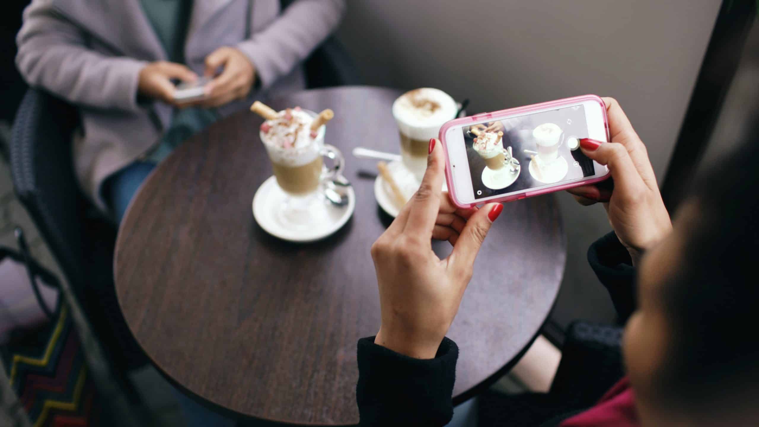 An overhead photograph taken from behind a person with red fingernails holding a smartphone with a pink case. The screen shows a live view of two gourmet coffee drinks on a round wooden table, which the person is about to photograph. In the background, another person is blurred, also using their phone. This visual captures the moment of creating authentic social media content for discovery.