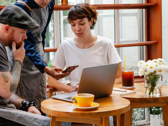 3 people sitting in a cafe looking at a computer and mobile phone thinking about their brand visibility, online reviews and Conversion Marketing brand sentiment reporting.
