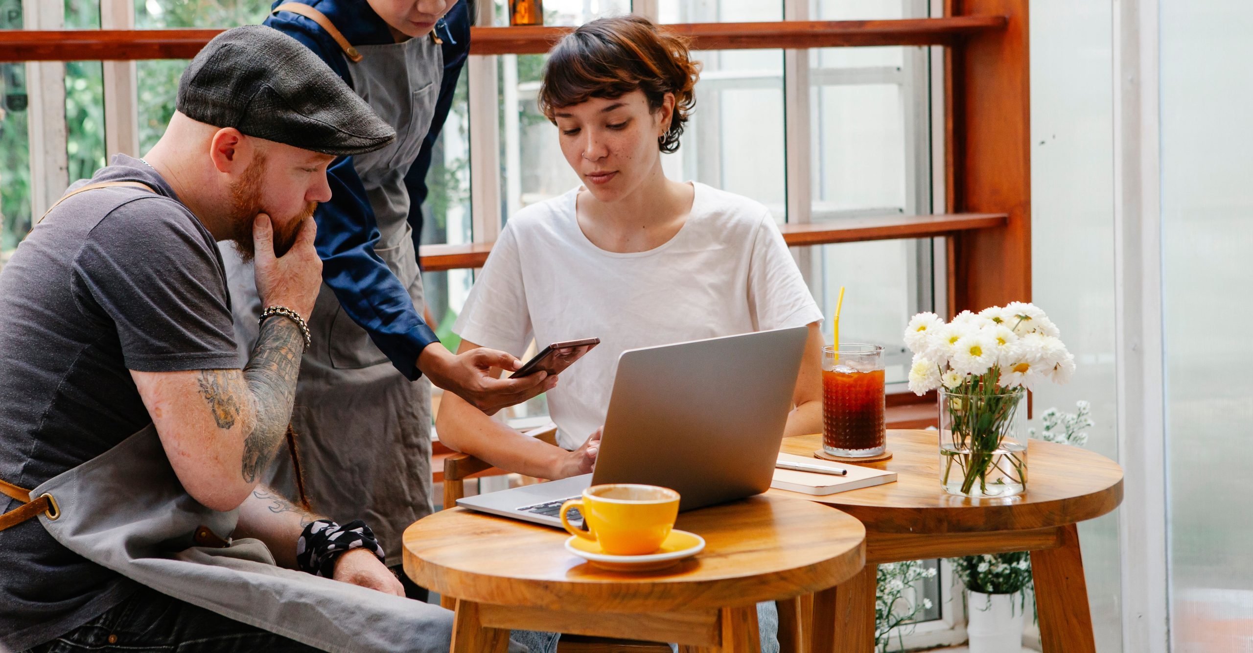 3 people sitting in a cafe looking at a computer and mobile phone thinking about their brand visibility, online reviews and Conversion Marketing brand sentiment reporting.